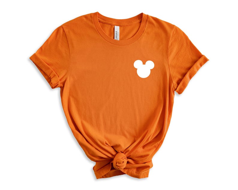 Puede incluir: Camiseta naranja de manga corta con una silueta blanca de Mickey Mouse en el pecho izquierdo.