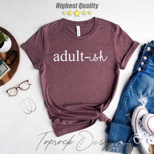May include: A maroon t-shirt with the text "adult-ish" in white lettering. The shirt is displayed on a white background with jeans, a camera, and a plant.