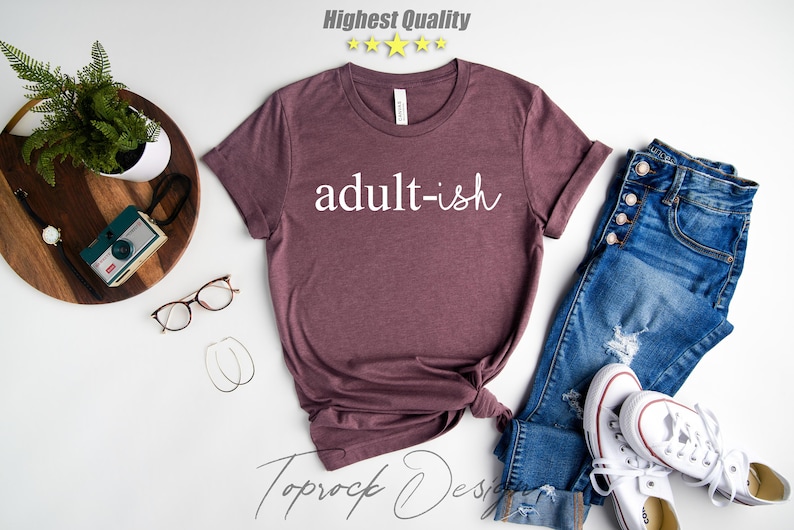 May include: A maroon t-shirt with the text "adult-ish" in white lettering. The shirt is displayed on a white background with jeans, a camera, and a plant.
