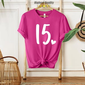 May include: A pink t-shirt with the number 15 printed in white on the front. The number is in a handwritten style with a small heart next to it.