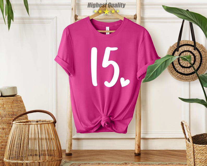 May include: A pink t-shirt with the number 15 printed in white on the front. The number is in a handwritten style with a small heart next to it.