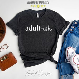 May include: A dark gray t-shirt with the text "adult-ish" in white lettering. The shirt is laid flat on a white surface with a pair of jeans, a brown purse, a pair of glasses, a potted succulent, and a vintage camera.