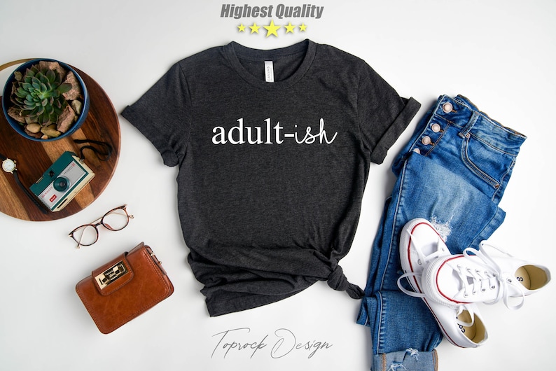 May include: A dark gray t-shirt with the text "adult-ish" in white lettering. The shirt is laid flat on a white surface with a pair of jeans, a brown purse, a pair of glasses, a potted succulent, and a vintage camera.
