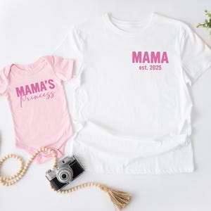 May include: A white t-shirt and a pink baby onesie. The t-shirt has the word "MAMA est. 2025" printed in pink. The onesie says "MAMA'S princess" in pink script. A vintage camera and decorative beads are also in the image.