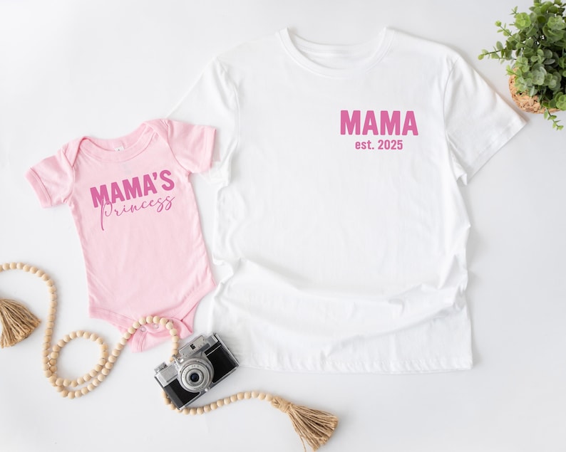 May include: A white t-shirt and a pink baby onesie. The t-shirt has the word "MAMA est. 2025" printed in pink. The onesie says "MAMA'S princess" in pink script. A vintage camera and decorative beads are also in the image.
