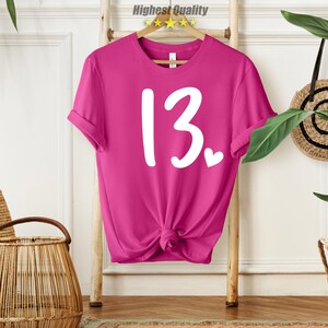 May include: Pink t-shirt with white text that says "13" with a small heart next to it.