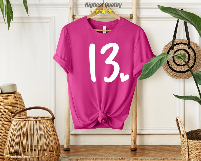 May include: Pink t-shirt with white text that says "13" with a small heart next to it.