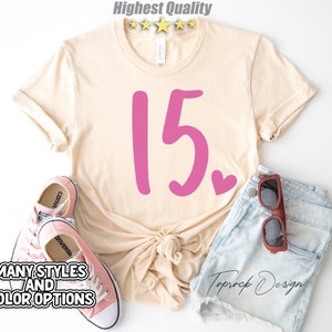 May include: A light beige t-shirt with a pink number 15 and a small pink heart. The shirt is tied in a knot at the front. The shirt is laid out on a white surface with a pair of pink Converse shoes and a pair of brown sunglasses on a pair of light blue denim shorts.