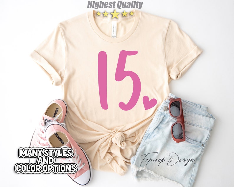 May include: A light beige t-shirt with a pink number 15 and a small pink heart. The shirt is tied in a knot at the front. The shirt is laid out on a white surface with a pair of pink Converse shoes and a pair of brown sunglasses on a pair of light blue denim shorts.