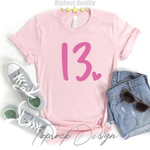 May include: Pink t-shirt with the number 13 and a small pink heart on a white background. The shirt is laying on a white surface with a pair of gray sneakers and a pair of denim shorts.