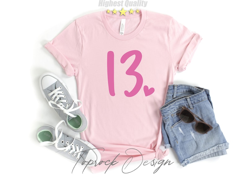 May include: Pink t-shirt with the number 13 and a small pink heart on a white background. The shirt is laying on a white surface with a pair of gray sneakers and a pair of denim shorts.