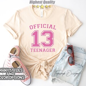 May include: A light beige t-shirt with the text "OFFICIAL 13 TEENAGER" in pink lettering. The number 13 is in a large, bold font with a white outline.