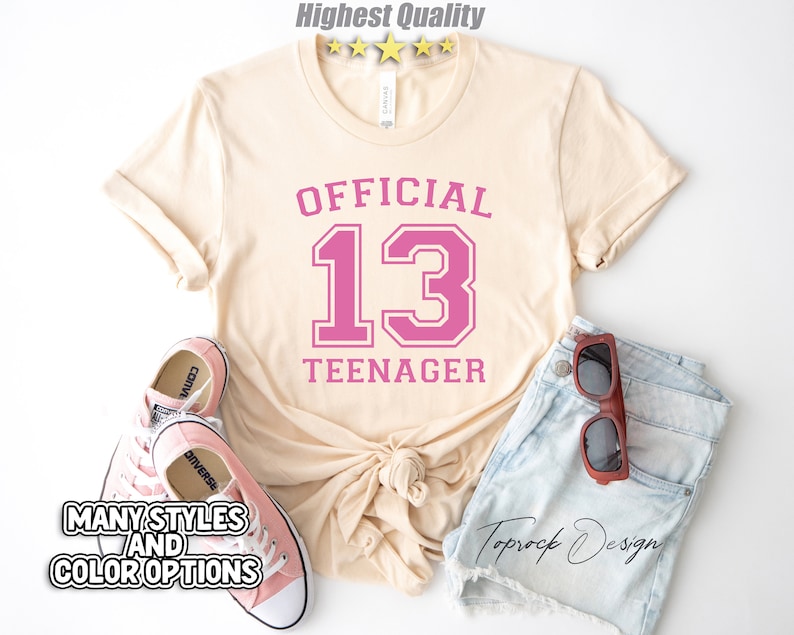 May include: A light beige t-shirt with the text "OFFICIAL 13 TEENAGER" in pink lettering. The number 13 is in a large, bold font with a white outline.