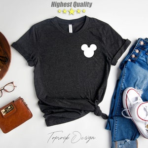 Puede incluir: Una camiseta gris oscuro con una silueta blanca de Mickey Mouse en la parte delantera. La camiseta est&aacute; colocada sobre una superficie blanca con un par de vaqueros, una cartera marr&oacute;n, unas gafas, una c&aacute;mara y una planta suculenta en maceta.