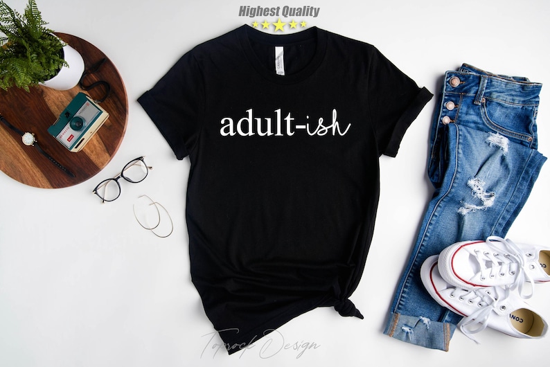 May include: Black t-shirt with white text that reads "adult-ish". The shirt is shown on a white background with a pair of jeans and sneakers.
