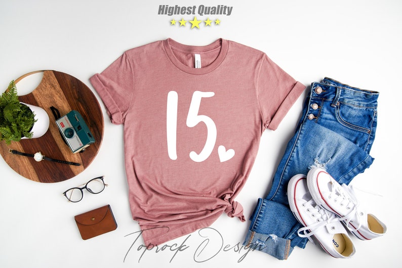 May include: A pink t-shirt with the number 15 and a small heart printed in white on the front. The shirt is laid flat on a white surface with a pair of jeans, a pair of white sneakers, a wooden cutting board, a potted plant, a camera, and a pair of glasses.