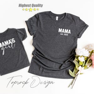 May include: Two dark gray t-shirts, one adult and one child size, with white text. The adult shirt says "MAMA est. 2025" and the child's shirt says "MAMA'S girl". A bouquet of white and pink flowers is to the right.