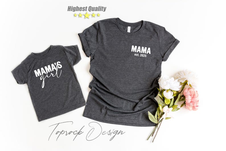 May include: Two dark gray t-shirts, one adult and one child size, with white text. The adult shirt says "MAMA est. 2025" and the child's shirt says "MAMA'S girl". A bouquet of white and pink flowers is to the right.