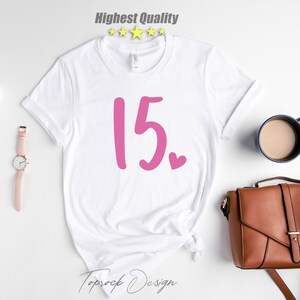 May include: White t-shirt with pink lettering that says "15."  There is a small pink heart next to the number.