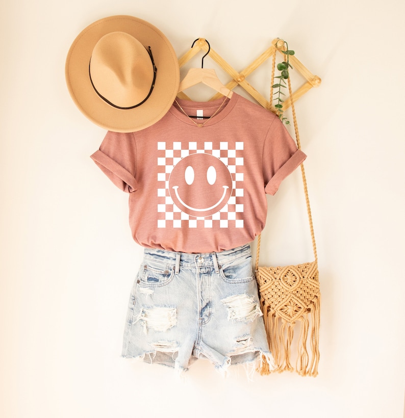 May include: A pink t-shirt with a white smiley face on a checkered background. The shirt is hanging on a wooden hanger and is paired with denim shorts and a straw bag.