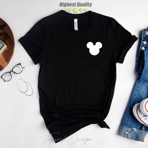 Puede incluir: Camiseta negra con un gr&aacute;fico de la cabeza de Mickey Mouse blanco en la parte delantera. La camiseta est&aacute; colocada sobre una superficie blanca con un par de jeans y zapatillas blancas.