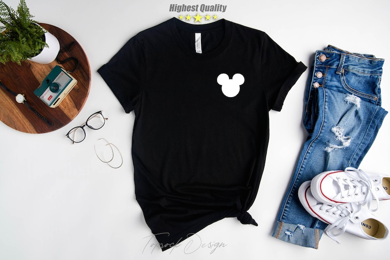 Puede incluir: Camiseta negra con un gr&aacute;fico de la cabeza de Mickey Mouse blanco en la parte delantera. La camiseta est&aacute; colocada sobre una superficie blanca con un par de jeans y zapatillas blancas.