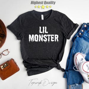 May include: A dark gray t-shirt with the text "Lil Monster" in white dripping letters. The shirt is laid flat on a white surface with a pair of jeans, a wallet, a camera, and a plant in a pot.