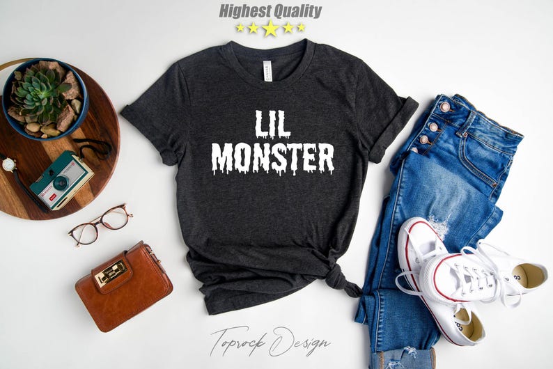 May include: A dark gray t-shirt with the text "Lil Monster" in white dripping letters. The shirt is laid flat on a white surface with a pair of jeans, a wallet, a camera, and a plant in a pot.