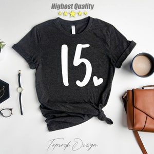 May include: A dark gray t-shirt with the number "15" and a small heart printed in white on the front. The shirt is laid flat on a white surface with other accessories.