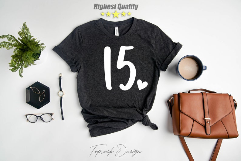 May include: A dark gray t-shirt with the number "15" and a small heart printed in white on the front. The shirt is laid flat on a white surface with other accessories.