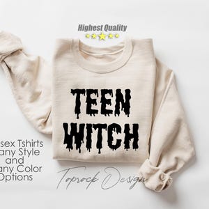 May include: A beige sweatshirt with the text "TEEN WITCH" in black dripping letters. The text is centered on the front of the sweatshirt. The sweatshirt is folded in half and laid flat on a white surface.