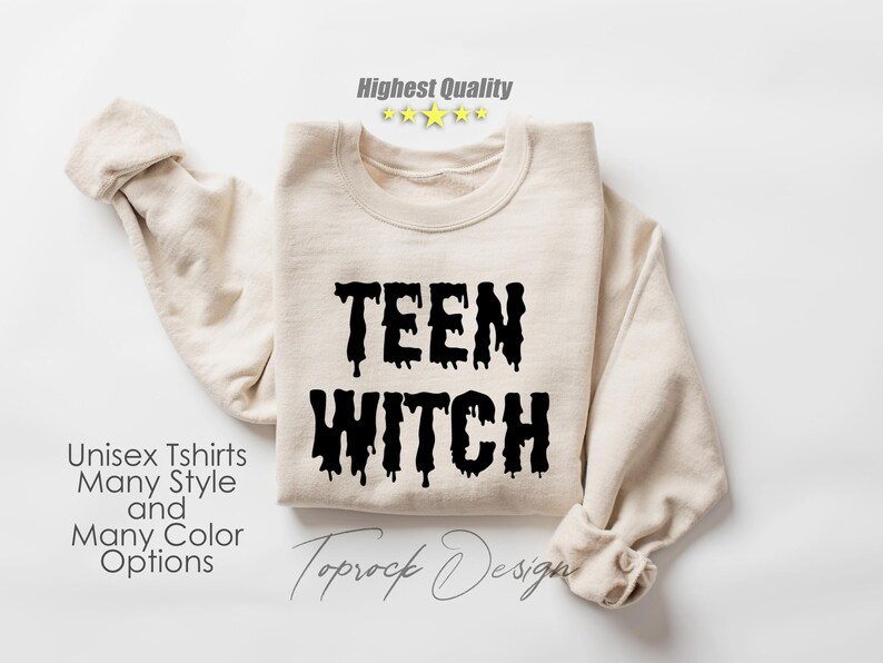 May include: A beige sweatshirt with the text "TEEN WITCH" in black dripping letters. The text is centered on the front of the sweatshirt. The sweatshirt is folded in half and laid flat on a white surface.