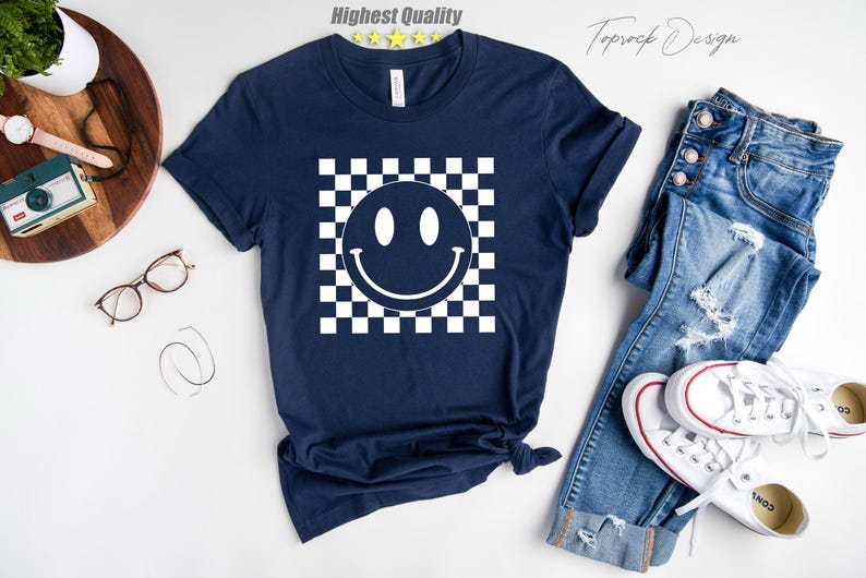 May include: A navy blue t-shirt with a white checkered pattern and a white smiley face in the center.
