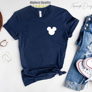 Puede incluir: Una camiseta azul marino con una silueta blanca de Mickey Mouse en la parte delantera. La camiseta est&aacute; colocada sobre una superficie blanca con un par de vaqueros y zapatillas blancas.