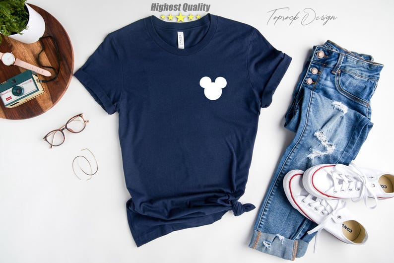 Puede incluir: Una camiseta azul marino con una silueta blanca de Mickey Mouse en la parte delantera. La camiseta est&aacute; colocada sobre una superficie blanca con un par de vaqueros y zapatillas blancas.