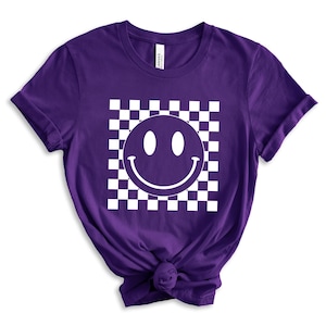 May include: Purple short-sleeved t-shirt with a white checkered pattern and a white smiley face graphic.