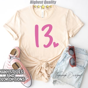 May include: A beige t-shirt with a pink number "13" and a small pink heart. The shirt is laid out on a white background with a pair of pink sneakers and denim shorts with brown sunglasses.