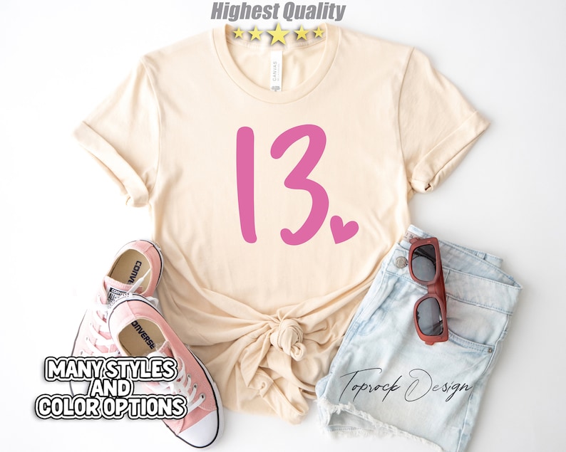 May include: A beige t-shirt with a pink number "13" and a small pink heart. The shirt is laid out on a white background with a pair of pink sneakers and denim shorts with brown sunglasses.