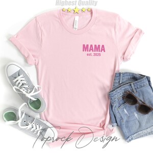 May include: A light pink t-shirt with the word "MAMA" and the text "est. 2025" printed in pink. The shirt is paired with denim shorts, sunglasses, and gray sneakers.
