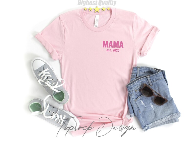 May include: A light pink t-shirt with the word "MAMA" and the text "est. 2025" printed in pink. The shirt is paired with denim shorts, sunglasses, and gray sneakers.