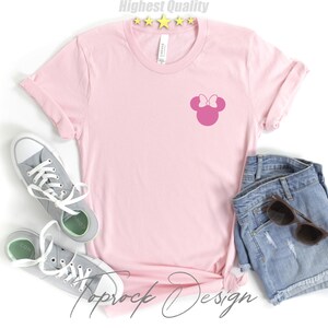 Puede incluir: Una camiseta rosa con una silueta rosa de la cabeza de Minnie Mouse en la parte delantera. La camiseta est&aacute; colocada sobre una superficie blanca con un par de zapatillas grises y unos pantalones cortos de mezclilla.
