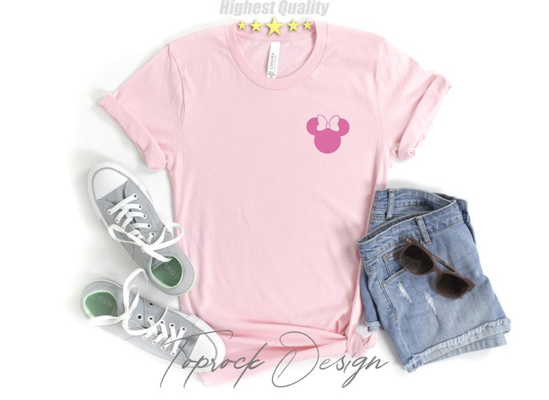 Puede incluir: Una camiseta rosa con una silueta rosa de la cabeza de Minnie Mouse en la parte delantera. La camiseta est&aacute; colocada sobre una superficie blanca con un par de zapatillas grises y unos pantalones cortos de mezclilla.