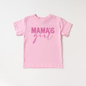May include: A light pink toddler t-shirt with the words "MAMA'S girl" printed in a darker pink script across the front. The shirt has short sleeves and a crew neck, perfect for casual wear.