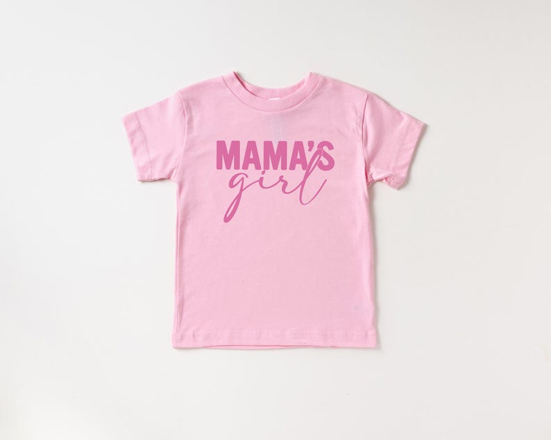 May include: A light pink toddler t-shirt with the words "MAMA'S girl" printed in a darker pink script across the front. The shirt has short sleeves and a crew neck, perfect for casual wear.