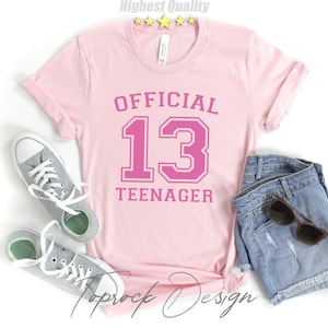 May include: Pink t-shirt with the text "OFFICIAL 13 TEENAGER" in a pink athletic font.