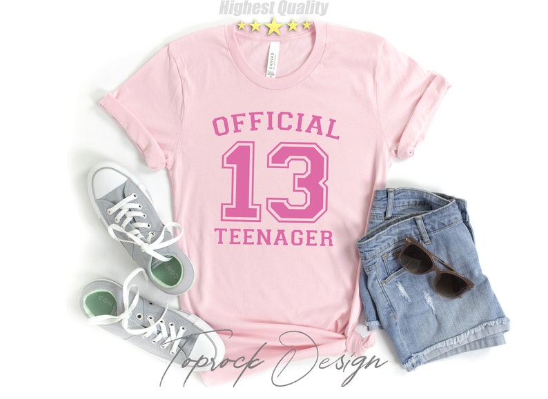 May include: Pink t-shirt with the text "OFFICIAL 13 TEENAGER" in a pink athletic font.