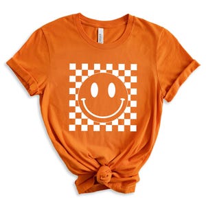 May include: Orange t-shirt with a white smiley face in a checkered pattern.