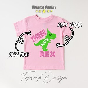 May include: Pink t-shirt with a green dinosaur wearing a party hat. The dinosaur says "Three Name Rex".