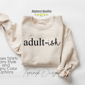 May include: A beige sweatshirt with the text "adult-ish" printed in black. The sweatshirt is folded and laid flat on a white surface. The text "Unisex Tshirts Many Style and Many Color Options" is printed below the sweatshirt.