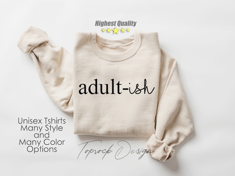 May include: A beige sweatshirt with the text "adult-ish" printed in black. The sweatshirt is folded and laid flat on a white surface. The text "Unisex Tshirts Many Style and Many Color Options" is printed below the sweatshirt.